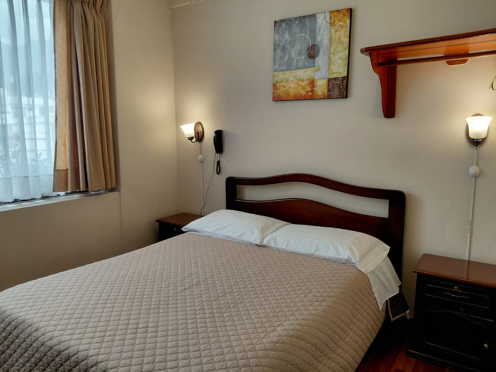Habitación simple con cama doble, cuadro artístico y repisa de madera en hostal Quito